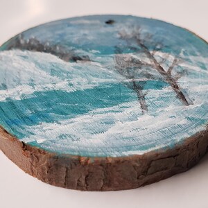 May include: Hand-painted wooden ornament depicting a winter landscape. The artwork shows a snowy scene with teal and white brushstrokes, and silhouetted trees. The round ornament has a natural wood edge and a small hole for hanging.