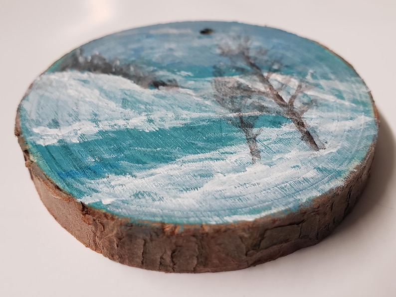 May include: Hand-painted wooden ornament depicting a winter landscape. The artwork shows a snowy scene with teal and white brushstrokes, and silhouetted trees. The round ornament has a natural wood edge and a small hole for hanging.