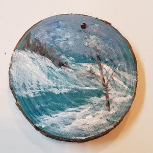 May include: A circular wooden slice featuring a painted seascape. The artwork shows a blue ocean with white waves, a dark cliff, and a tree. The painting uses shades of blue, white, and brown.