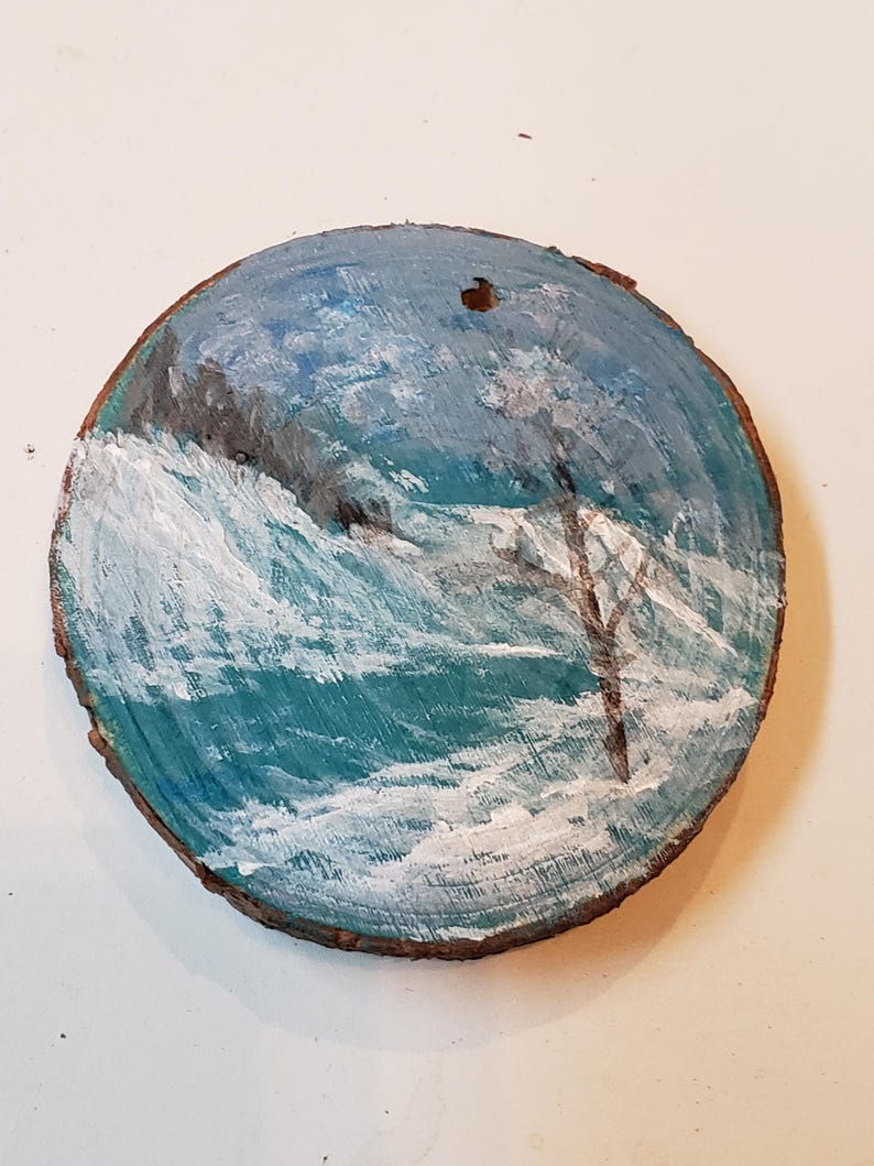 May include: A circular wooden slice featuring a painted seascape. The artwork shows a blue ocean with white waves, a dark cliff, and a tree. The painting uses shades of blue, white, and brown.