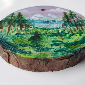 May include: A circular, hand-painted wooden slice with a landscape scene. The artwork shows a lake surrounded by green fields and trees, under a purple and blue sky. The wood has a natural, brown bark edge.