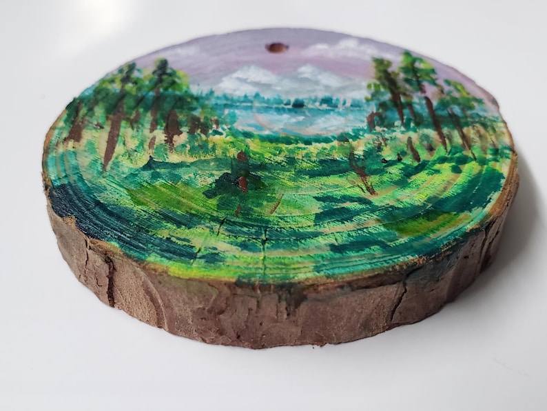 May include: A circular, hand-painted wooden slice with a landscape scene. The artwork shows a lake surrounded by green fields and trees, under a purple and blue sky. The wood has a natural, brown bark edge.