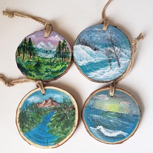May include: Four circular wooden ornaments, each hand-painted with a different landscape scene. The scenes depict mountains, forests, and water, using various colours. Each has a twine loop for hanging, perfect for a rustic touch.
