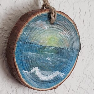 May include: A circular wooden slice featuring a painted seascape. The artwork shows a blue ocean with white waves, a yellow and green sunset, and a hanging rope. The wood has a natural, brown bark edge.