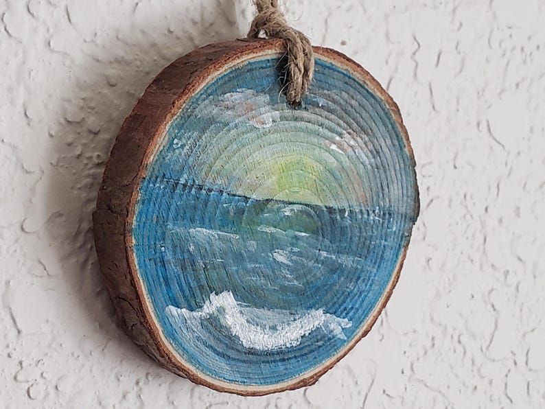 May include: A circular wooden slice featuring a painted seascape. The artwork shows a blue ocean with white waves, a yellow and green sunset, and a hanging rope. The wood has a natural, brown bark edge.