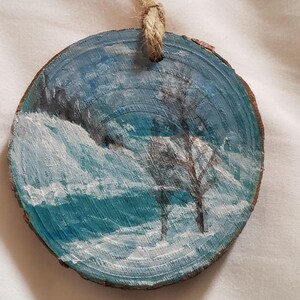 May include: Round, hand-painted wooden ornament with a winter scene. The artwork shows a snowy field, a bare tree, and a blue sky. A natural twine loop is attached for hanging. The ornament is made from a slice of wood.