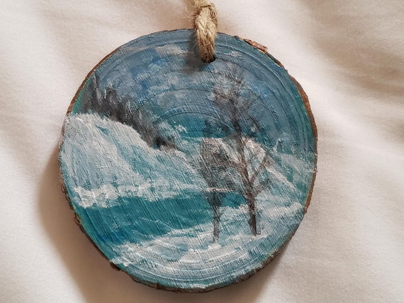 May include: Round, hand-painted wooden ornament with a winter scene. The artwork shows a snowy field, a bare tree, and a blue sky. A natural twine loop is attached for hanging. The ornament is made from a slice of wood.