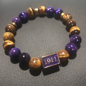 May include: A beaded bracelet featuring alternating purple and brown tiger eye beads. A rectangular charm with the number "1911" in white lettering is centered on the bracelet. The bracelet is made of natural stone beads.