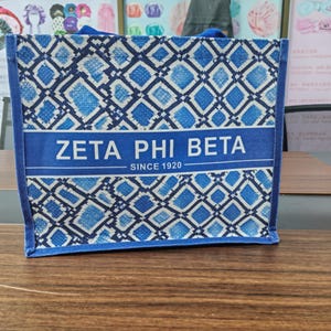 May include: A rectangular blue and white tote bag featuring a repeating diamond pattern. A central horizontal band displays the text ZETA PHI BETA in white capital letters, with the text SINCE 1920 positioned directly below it in a smaller font.