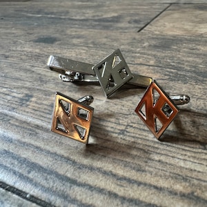 May include: Three silver cufflinks with geometric designs. The cufflinks are in the shape of a diamond with a geometric pattern inside. The cufflinks are on a wooden surface.