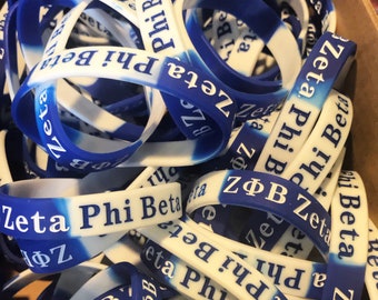Puede incluir: Un montón de pulseras de goma azul y blanca con el texto "Zeta Phi Beta" impreso en ellas.