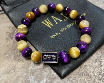 May include: A beaded bracelet featuring alternating purple and gold-colored tiger eye stones. The bracelet is displayed on a black leather pouch with the text "WAJ" and "shopwaj.com". A small, square, blue charm with text is also visible.