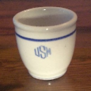 May include: A small white ceramic cup with a blue band and the letters "USH" in blue.
