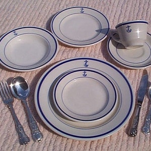 May include: A nautical-themed white porcelain dinnerware set with blue trim and anchor details. The set includes plates, bowls, a teacup and saucer, and silverware. The plates and bowls have a blue border and an anchor symbol.