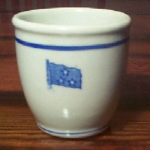 May include: A white ceramic cup featuring a blue horizontal band near the rim and a blue base. The exterior surface displays a small blue rectangular flag graphic with three white stars arranged in a triangular pattern on the front side.