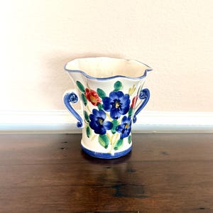 May include: A cream-colored ceramic vase with a scalloped rim and two blue scroll-shaped handles. The exterior features hand-painted blue flowers with green leaves and a single red flower, finished with a blue painted band around the base.