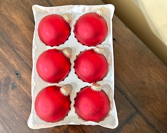 May include: Six red Christmas ornaments in a white cardboard container. Each ornament is round with a matte finish and a gold-colored cap. The ornaments are arranged in two rows of three.