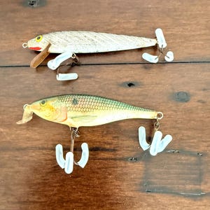 May include: Two vintage fishing lures. The top lure is silver with a brown fin and red accents. The bottom lure is green and yellow with a brown fin. Both lures have white plastic hooks and are on a wooden surface.