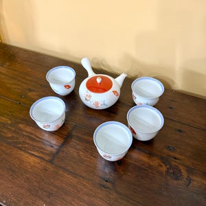 May include: A white ceramic tea set with a red lid and floral designs. The set includes a teapot and six small cups with blue rims and matching floral patterns. The tea set is arranged on a dark wooden surface.