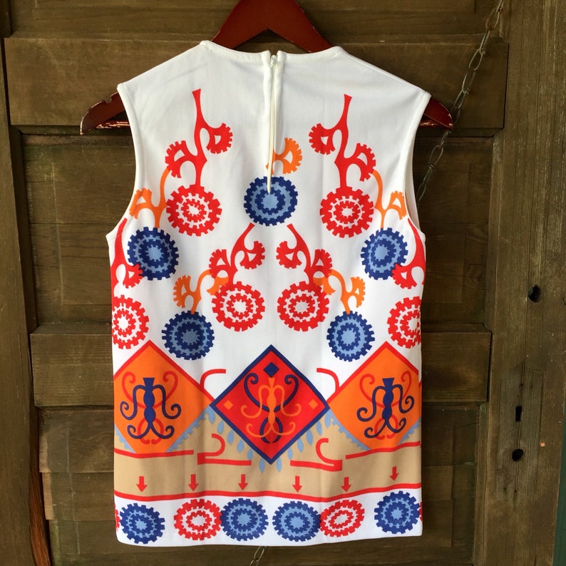 Puede incluir: Top sin mangas blanco con un vibrante estampado geom&eacute;trico en rojo, naranja, azul y beige. El dise&ntilde;o presenta formas florales y de diamante, con cierre de cremallera en la espalda. El top est&aacute; colgado de una percha de madera.
