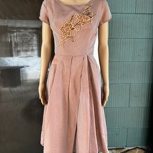 Könnte beinhalten: Staubrosa Kleid mit kurzen Ärmeln und knielangem Rock. Das Kleid hat ein dekoratives Blumendesign in Braun- und Goldtönen auf dem Oberteil. Das Kleid wird auf einer Schaufenquin ausgestellt.