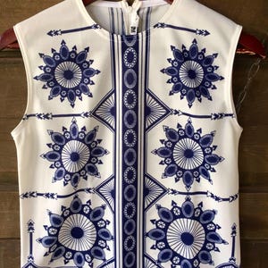 May include: White sleeveless top with a blue and white geometric pattern. The pattern features repeating circles, arrows, and diamond shapes.