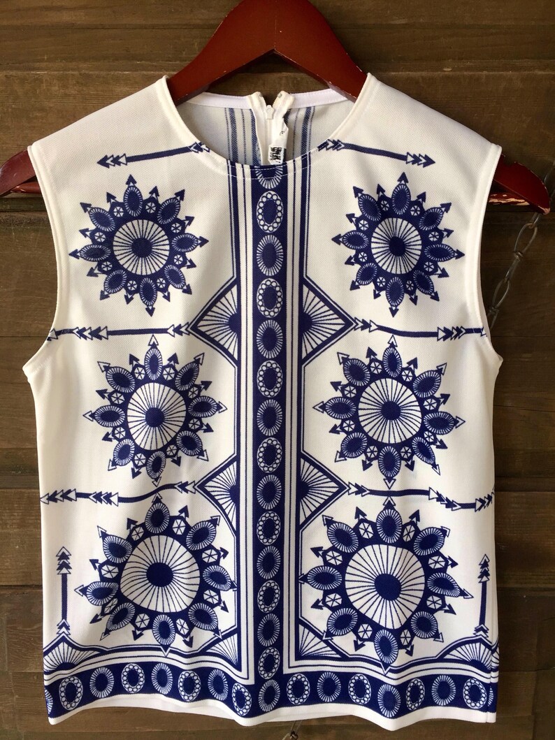 May include: White sleeveless top with a blue and white geometric pattern. The pattern features repeating circles, arrows, and diamond shapes.