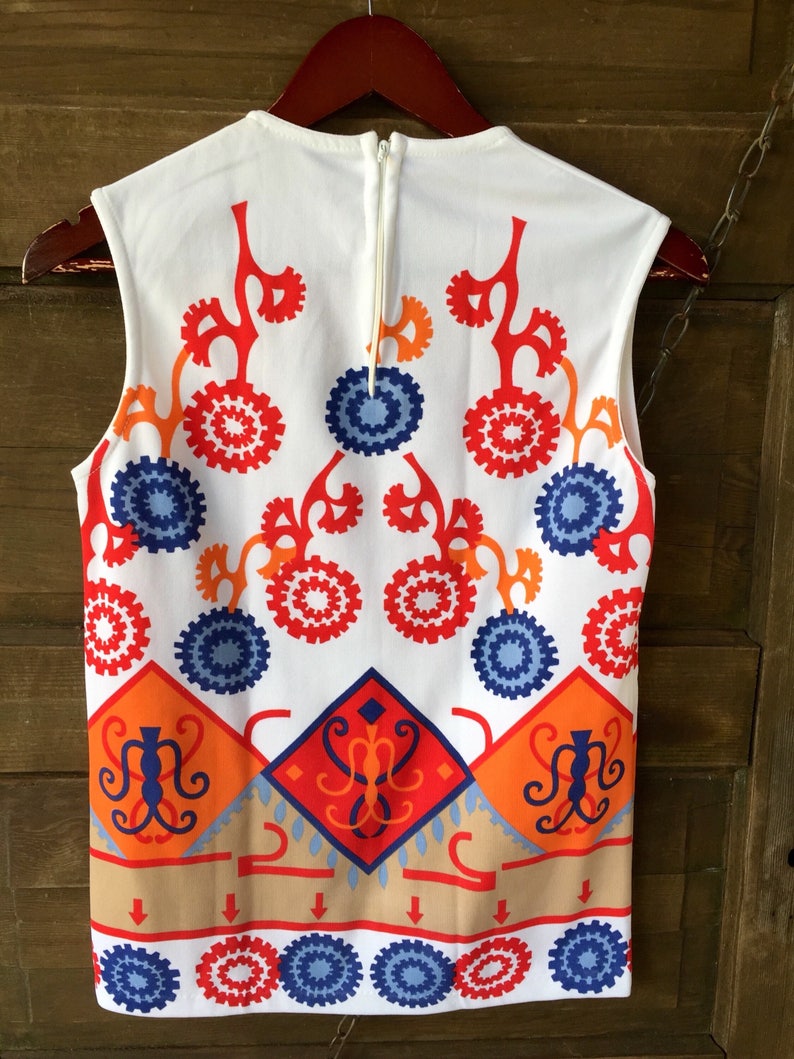 Puede incluir: Top sin mangas blanco con un vibrante estampado geom&eacute;trico en rojo, naranja, azul y beige. El dise&ntilde;o incluye formas florales y de diamante estilizadas. El top tiene una cremallera en la espalda y est&aacute; colgado de una percha de madera.