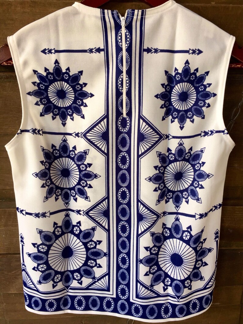 May include: White sleeveless top with a blue and white geometric pattern. The pattern features a repeating design of circles, arrows, and stars.