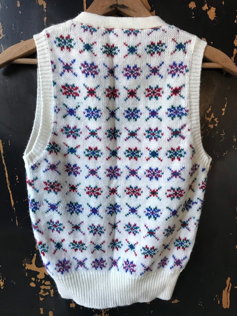 May include: White sleeveless sweater vest with a geometric pattern in red, blue, and green.