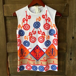 Puede incluir: Top sin mangas blanco con un llamativo estampado geom&eacute;trico en rojo, naranja y azul. El dise&ntilde;o incluye formas circulares y de diamante, creando una est&eacute;tica retro. El top est&aacute; colgado de una percha de madera.