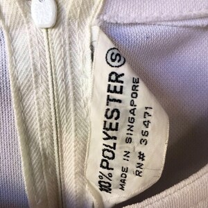 Puede incluir: Primer plano de una prenda de color crema con cremallera y bot&oacute;n blanco en el escote. Una etiqueta dice "100% POLYESTER" y "MADE IN SINGAPORE". La prenda parece ser vintage.