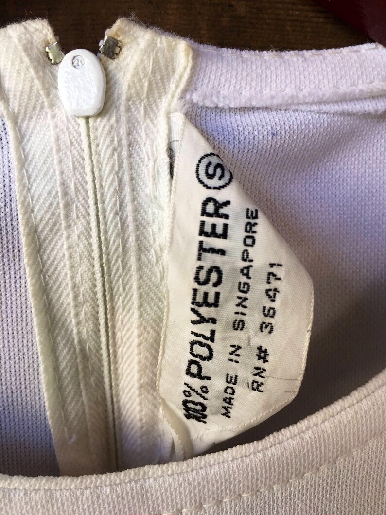 Puede incluir: Primer plano de una prenda de color crema con cremallera y bot&oacute;n blanco en el escote. Una etiqueta dice "100% POLYESTER" y "MADE IN SINGAPORE". La prenda parece ser vintage.