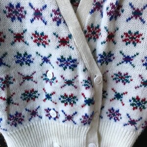 May include: White cardigan with a colorful geometric pattern. The cardigan has a V-neck and a button closure.