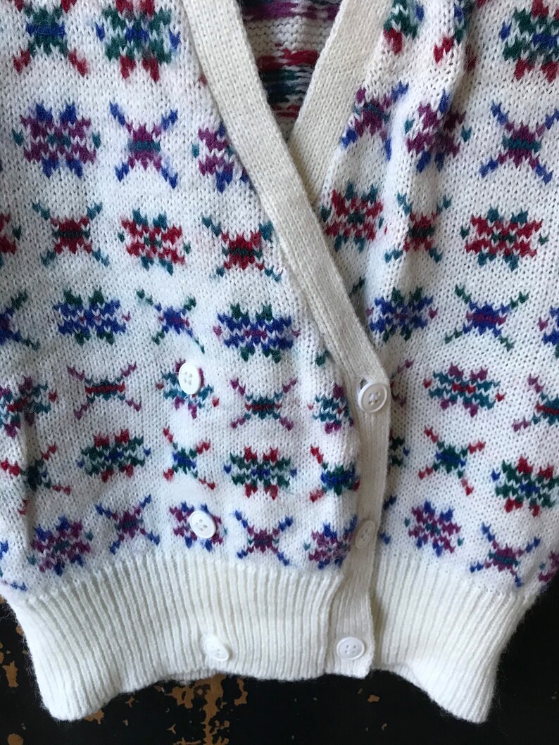 May include: White cardigan with a colorful geometric pattern. The cardigan has a V-neck and a button closure.