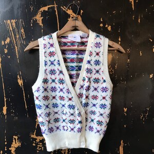 May include: White knitted vest with a colorful geometric pattern. The vest has a V-neck and button closure.