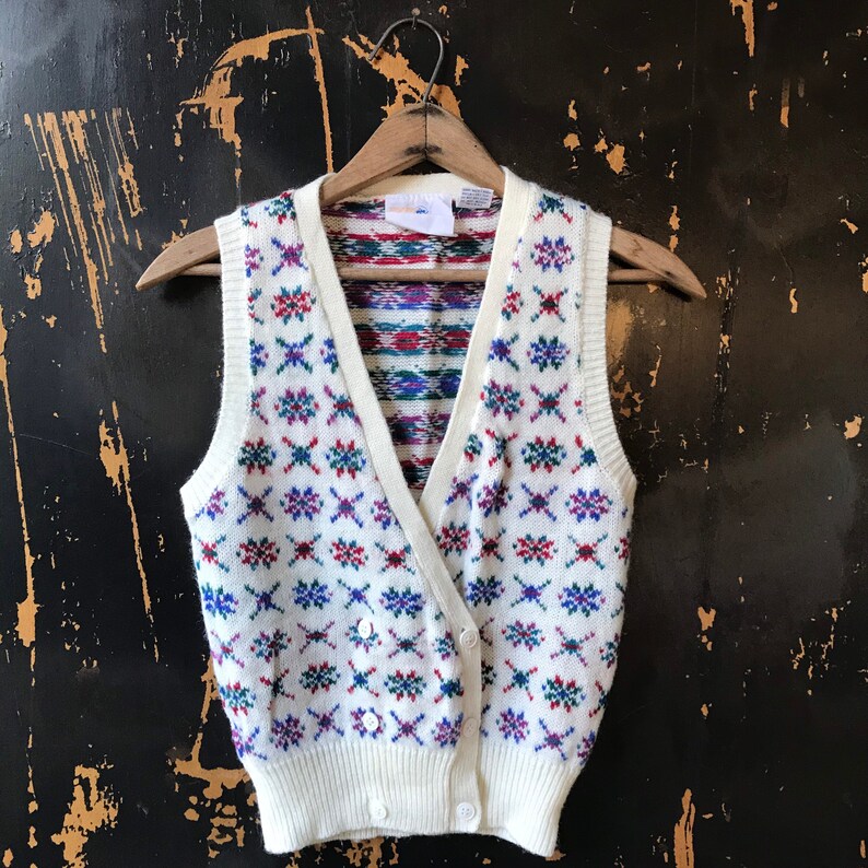 May include: White knitted vest with a colorful geometric pattern. The vest has a V-neck and button closure.