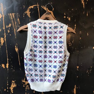 May include: White sleeveless sweater vest with a geometric pattern in red, blue, and green.