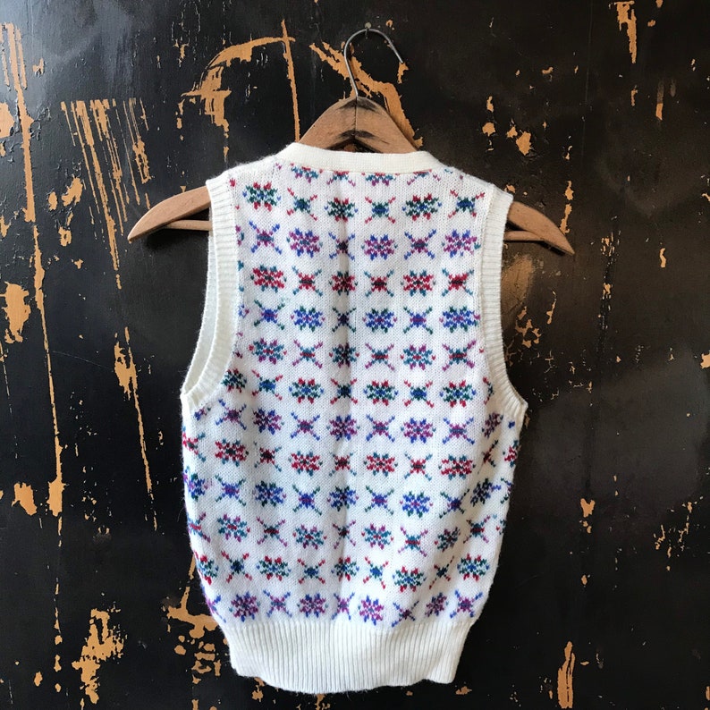 May include: White sleeveless sweater vest with a geometric pattern in red, blue, and green.