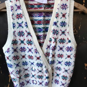 May include: White knitted vest with a geometric pattern in shades of red, blue, and green. The vest has a V-neck and button closure.