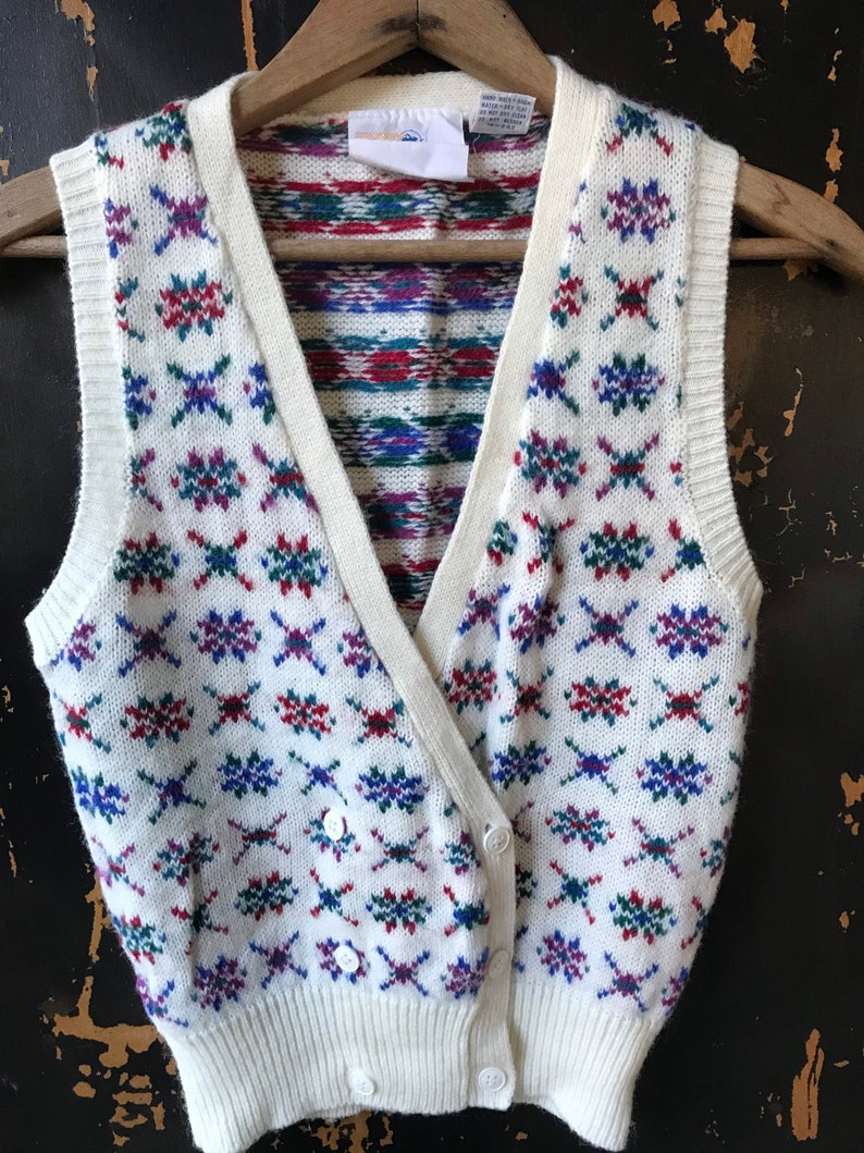 May include: White knitted vest with a geometric pattern in shades of red, blue, and green. The vest has a V-neck and button closure.