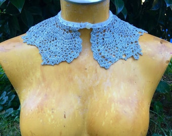 May include: A silver crochet collar with a pearl button closure. The collar is displayed on a yellow mannequin.