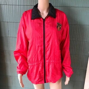 May include: A red nylon jacket with a black zipper and a black collar. The jacket has a small embroidered patch on the chest with a shield and a lion.