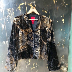 May include: A black velvet jacket with a gold paisley pattern and a red tag. The jacket has a button closure and long sleeves.