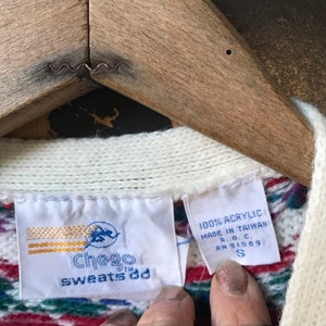 May include: White sweater with a multi-colored striped pattern. The sweater has a tag that reads "Chego sweats ad" and another tag that reads "100% Acrylic Made in Taiwan R.O.C. RN 51589 S".