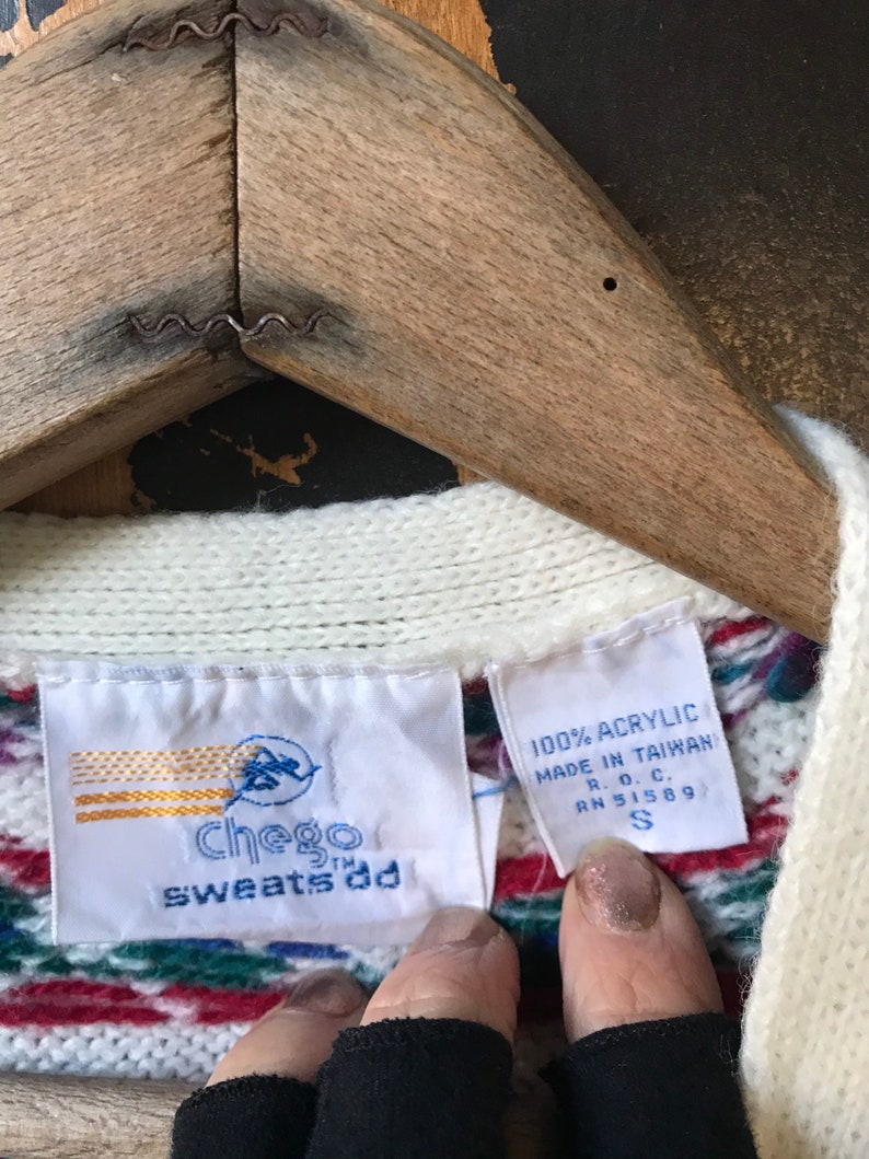 May include: White sweater with a multi-colored striped pattern. The sweater has a tag that reads "Chego sweats ad" and another tag that reads "100% Acrylic Made in Taiwan R.O.C. RN 51589 S".