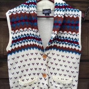 May include: A white knitted vest with a brown, blue, and red patterned collar. The vest has two pockets and a button closure. The vest has the brand name "Gitano" on the label.