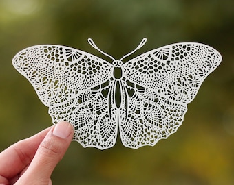 May include: White paper butterfly with intricate cut-out details, resembling lace. The butterfly is held in a hand against a blurred green background.