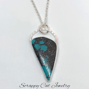 May include: A silver chain necklace with a turquoise pendant. The pendant is a teardrop shape with a silver ring around it. The pendant is set in a silver frame with a textured edge. The necklace is on a white background. "Scrappy Cat Jewelry" is written in cursive at the bottom of the image.