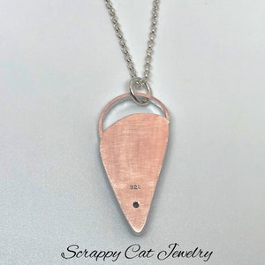 May include: A silver necklace with a teardrop-shaped pendant. The pendant has a cutout design and is marked with "925". The necklace is from "Scrappy Cat Jewelry".